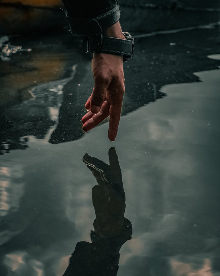 A hand reaches towards its reflection in a gloomy water puddle, creating a moody scene.