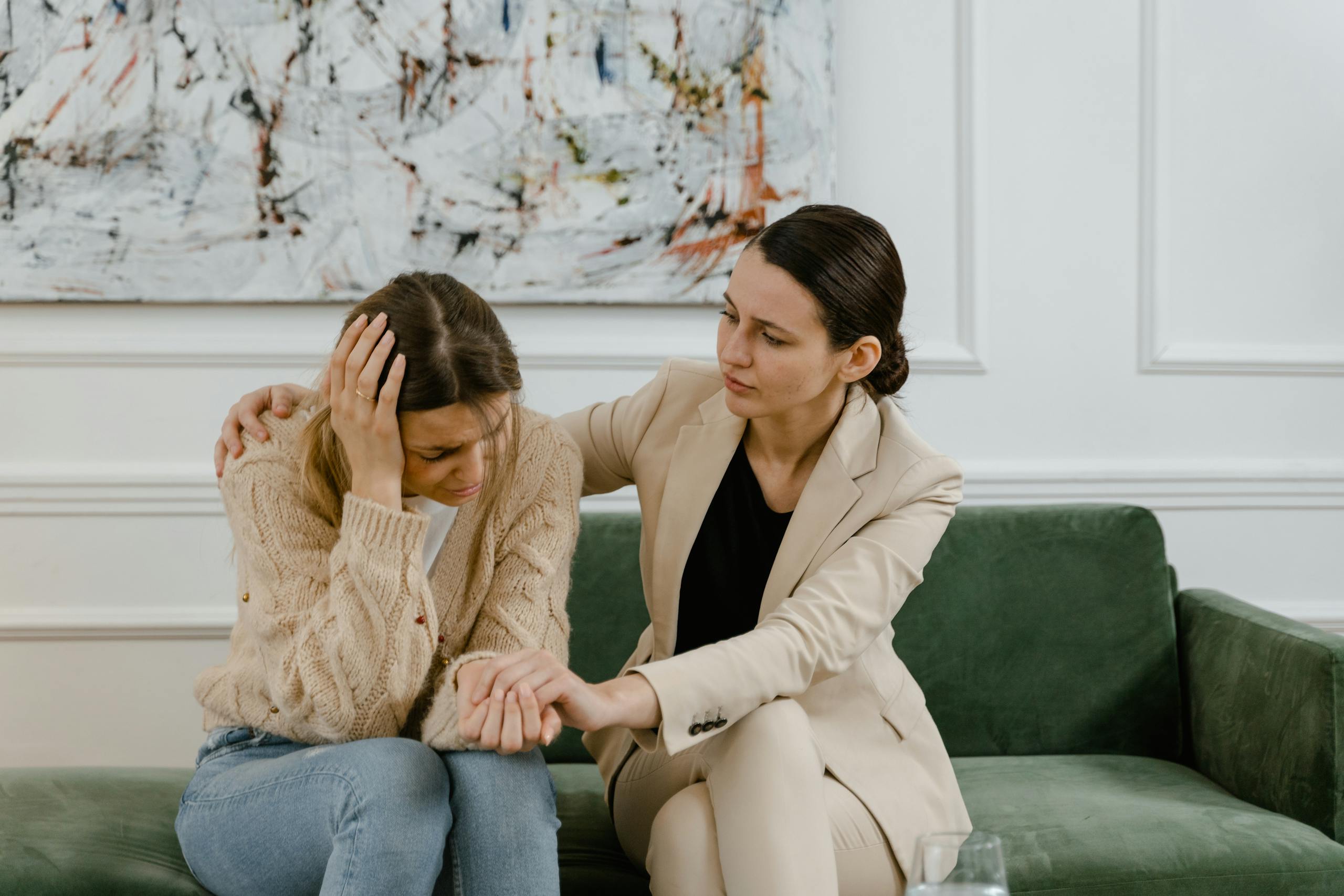 A woman comforts a friend in distress on a green couch indoors.