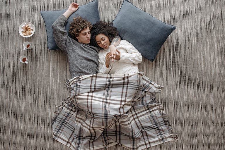 Interracial couple cuddling on the floor under a plaid blanket, enjoying a warm and cozy moment.